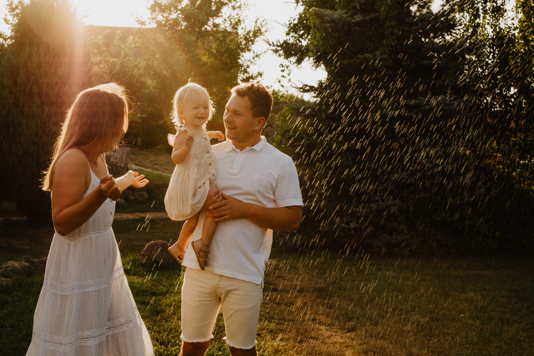 Uśmiechnięta rodzina w letnich ubraniach bawi się w ogrodzie w strugach wody w promieniach zachodzącego słońca. Mama, tata i mała córeczka.