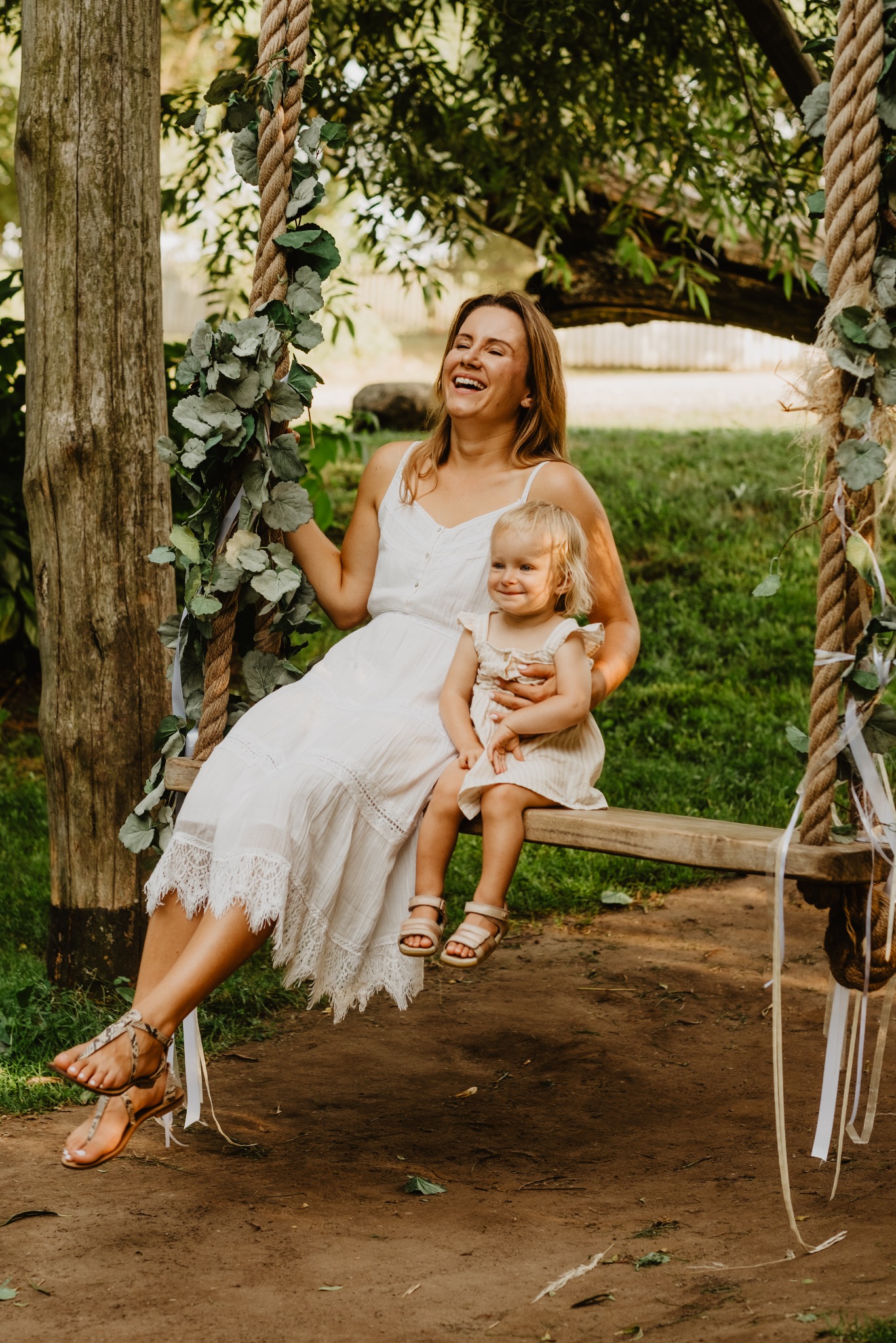 Uśmiechnięta mama w białej sukience siedzi na huśtawce z córeczką na kolanach. Huśtawka ozdobiona zielonymi liśćmi, naturalne światło, zielone tło.