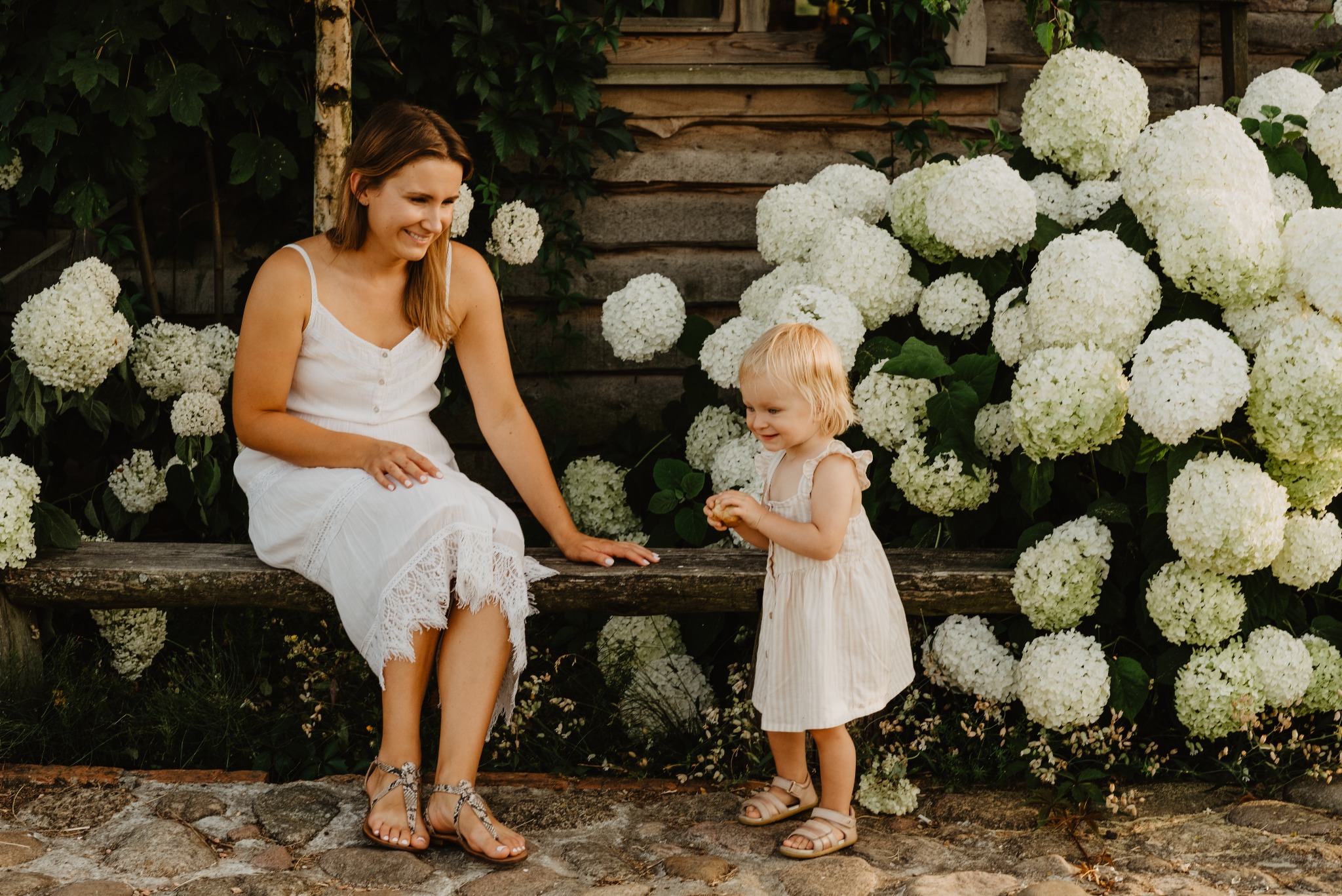 Mama w białej sukience siedzi na ławce obok córki w ogrodzie pełnym białych hortensji. Naturalne światło, ciepła tonacja, sesja w plenerze.
