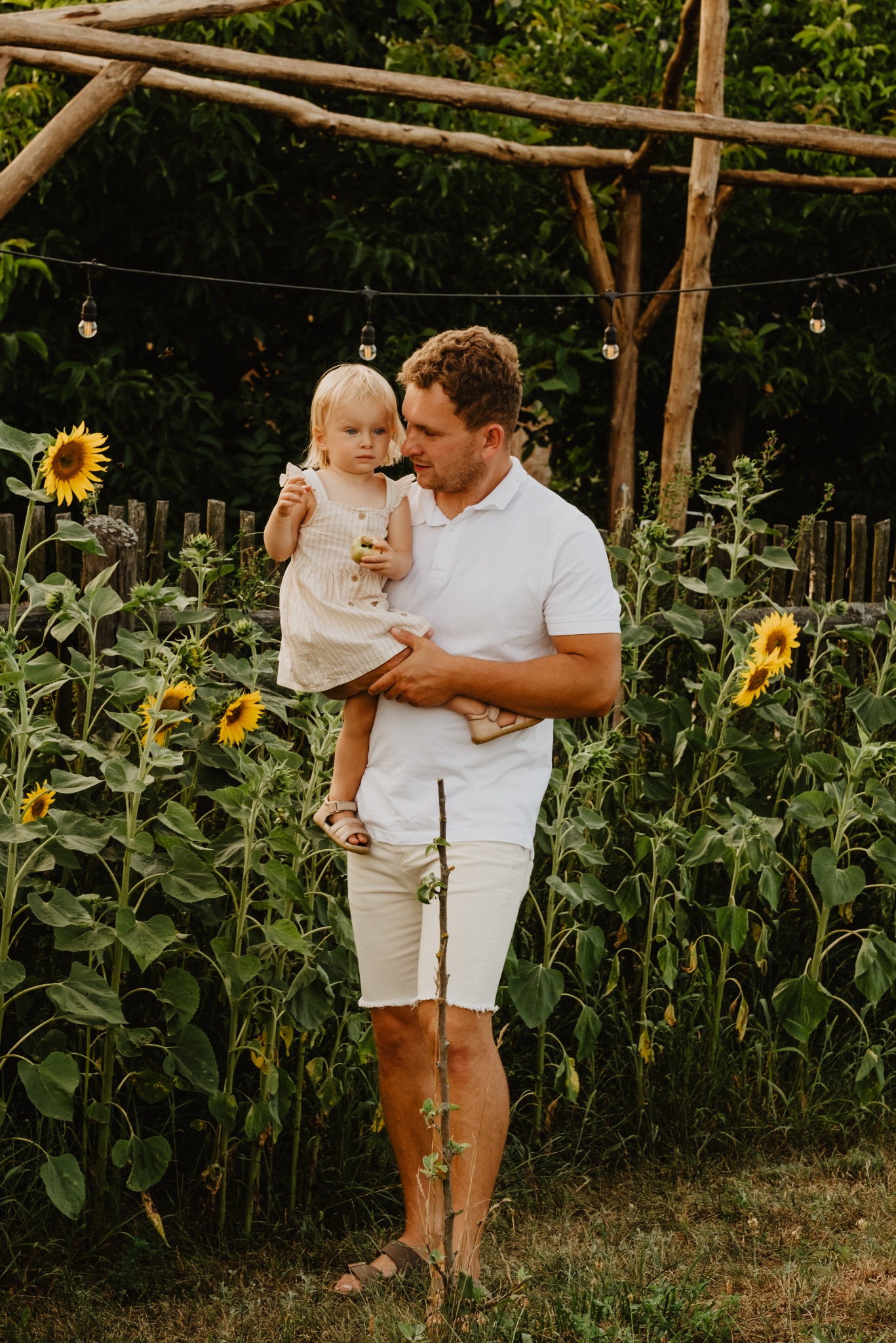 Ojciec trzyma córkę na tle słoneczników i drewnianej konstrukcji z żarówkami, uchwycone w ciepłym świetle podczas sesji rodzinnej.