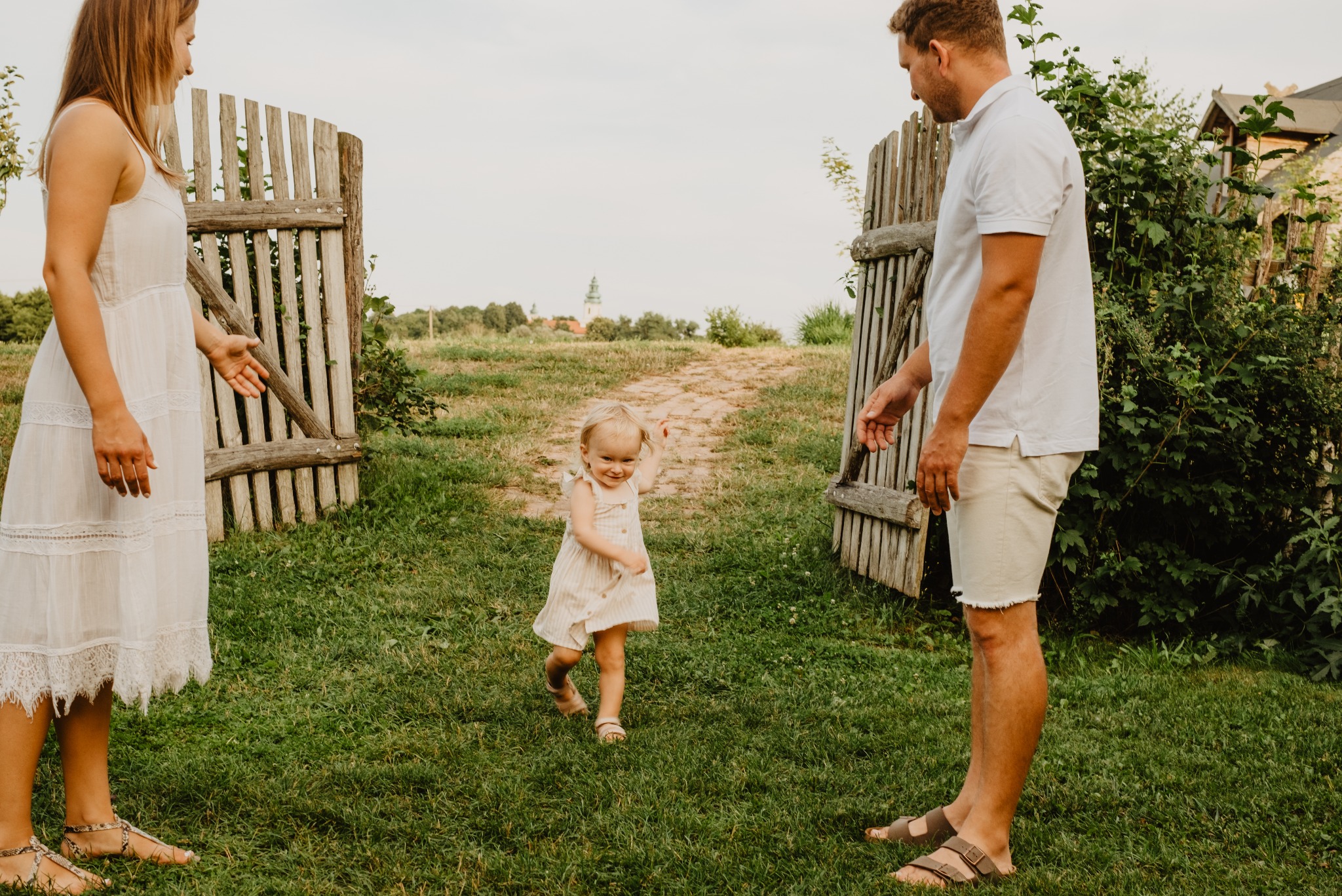 Uśmiechnięta dziewczynka biegnie po trawie w kierunku rodziców, którzy stoją przy drewnianej furtce. W tle widać budynki i zieleń. Naturalne światło, ciepła kolorystyka.