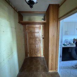 Kacper Dec - Korytarz przed remontem: ściany z widocznymi śladami, drewniana boazeria, stara podłoga. Widoczne wejście do kuchni. Lampa sufitowa w stylu retro.