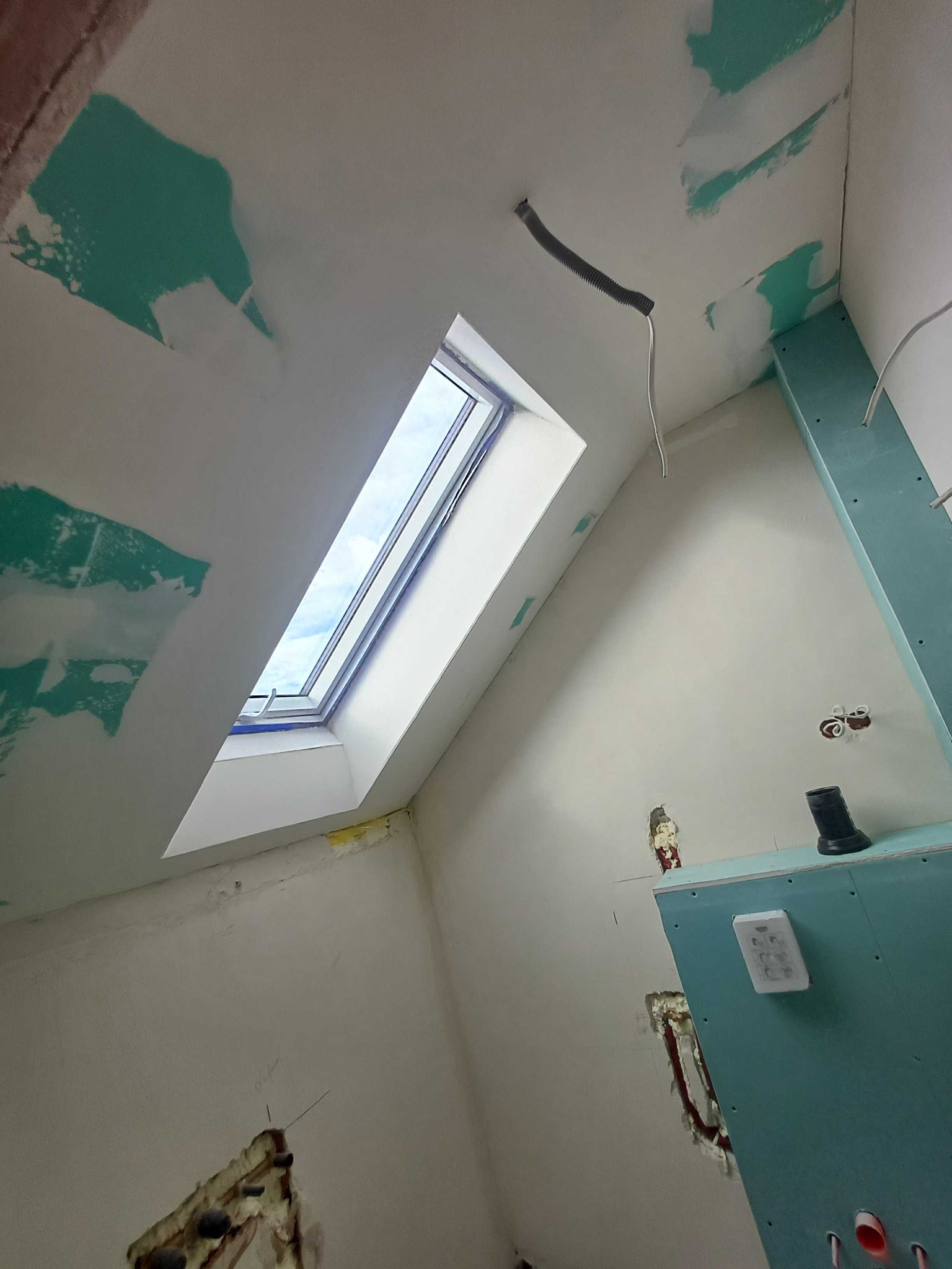 Poddasze w trakcie prac wykończeniowych: okno dachowe, ściany z płyt gipsowych, częściowo wyszpachlowane, widoczne instalacje elektryczne. Ujęcie z dołu.