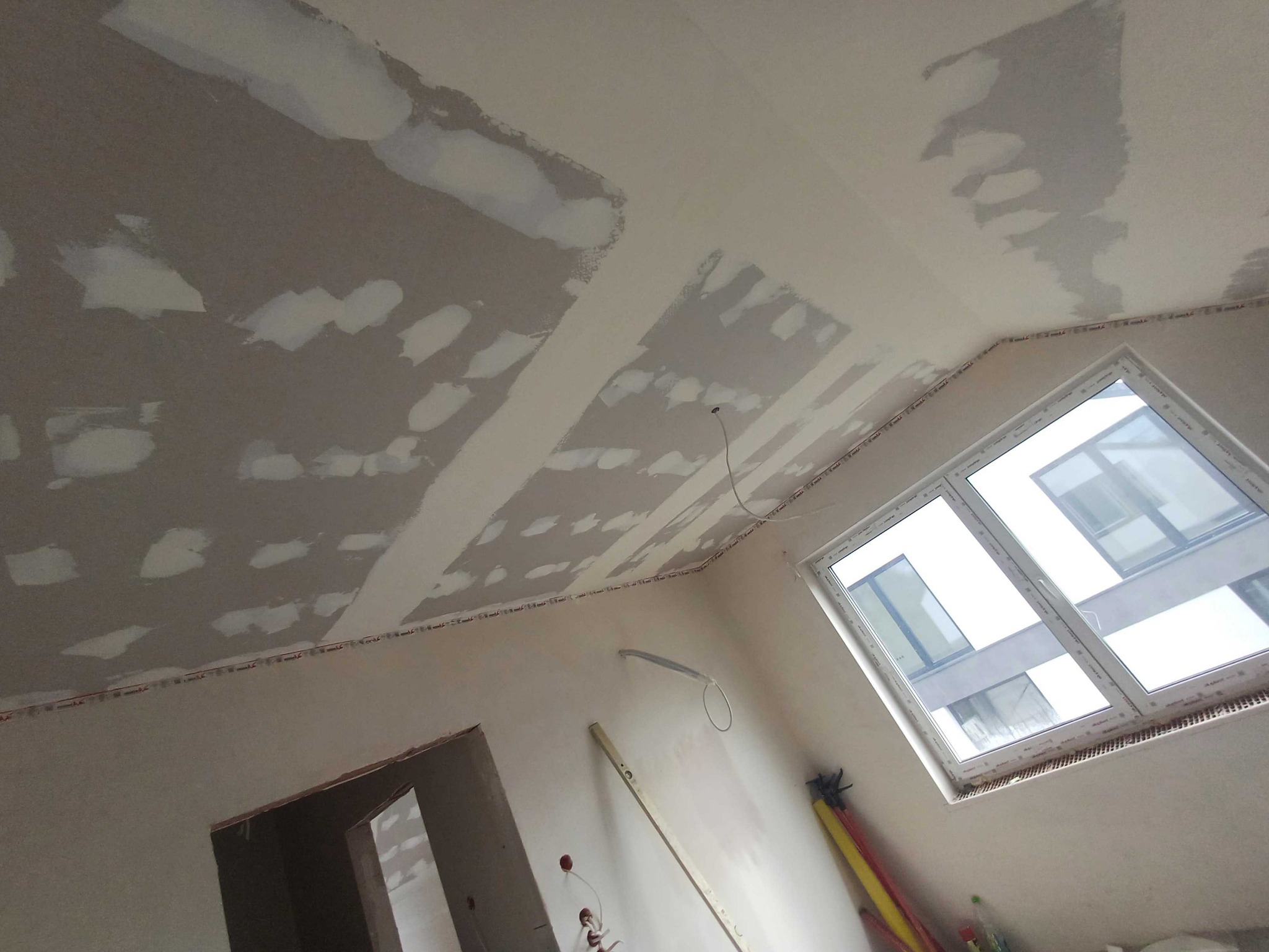 Wnętrze w trakcie szpachlowania: sufit i ściany z widocznymi łatami gładzi, okno dachowe, poziomica oparta o ścianę. Prace wykończeniowe w nowym budownictwie.