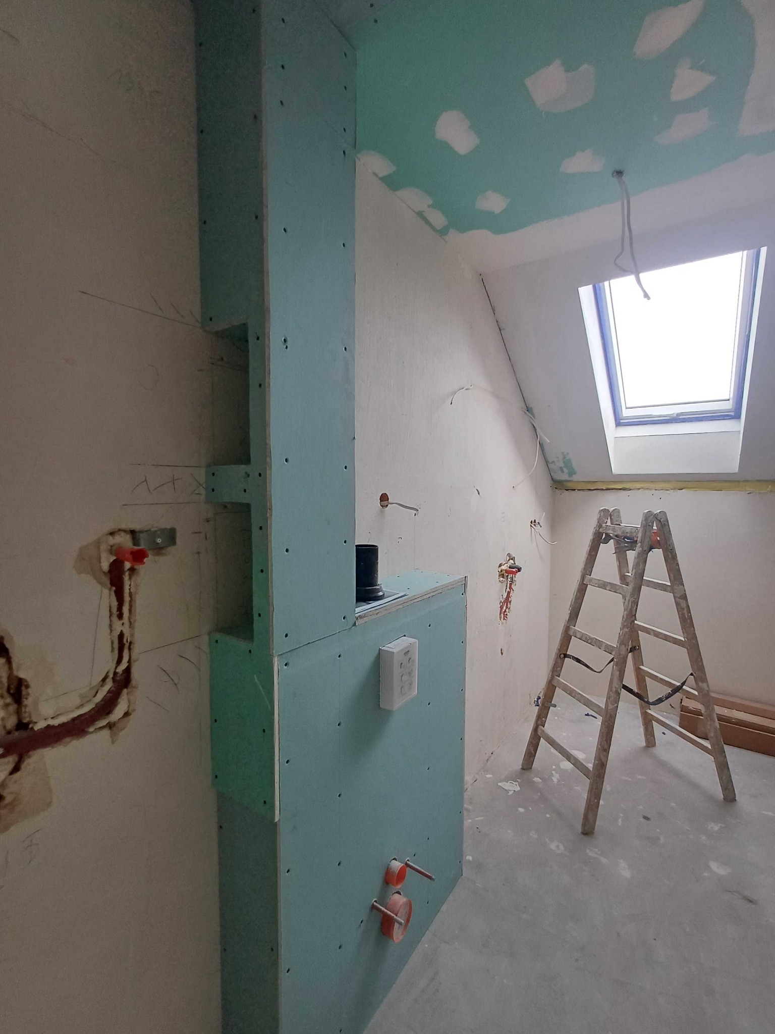 Poddasze w trakcie prac: zabudowa z płyty g-k, instalacje, drabina. Sufit z widocznymi łatami szpachli. Okno dachowe wpuszcza naturalne światło.