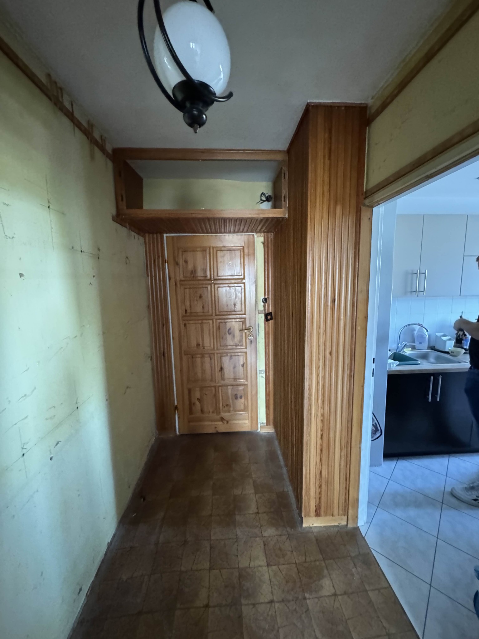 Korytarz przed remontem: ściany z widocznymi śladami, drewniana boazeria, stara podłoga. Widoczne wejście do kuchni. Lampa sufitowa w stylu retro.