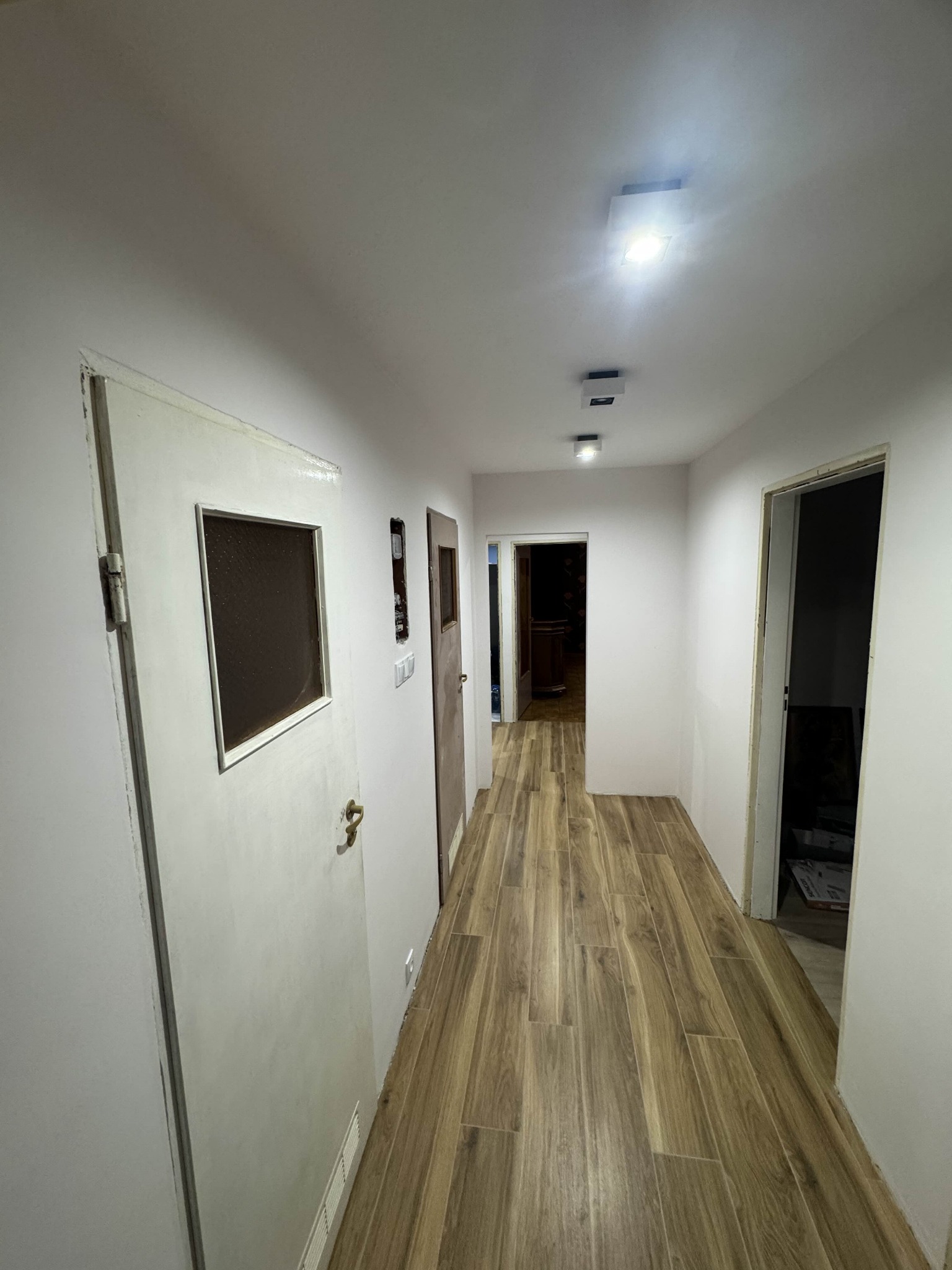 Korytarz po remoncie: drewnopodobna podłoga, białe ściany, nowe oświetlenie sufitowe. Widoczne drzwi z szybą i otwory drzwiowe do innych pomieszczeń.