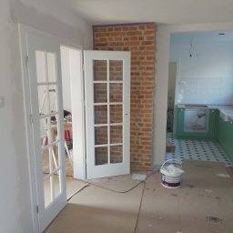 damian irla - Wnętrze w trakcie prac: białe, przeszklone drzwi uchylne, fragment ściany z cegły zabezpieczony taśmą, podłoga wyłożona kartonem, widoczne narzędzia malarskie.