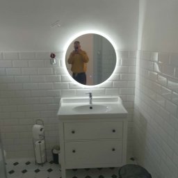 damian irla - Biała łazienka z okrągłym lustrem LED, białą umywalką z szafką, białe płytki na ścianach i czarno-biała podłoga. W lustrze odbija się osoba robiąca zdjęcie.