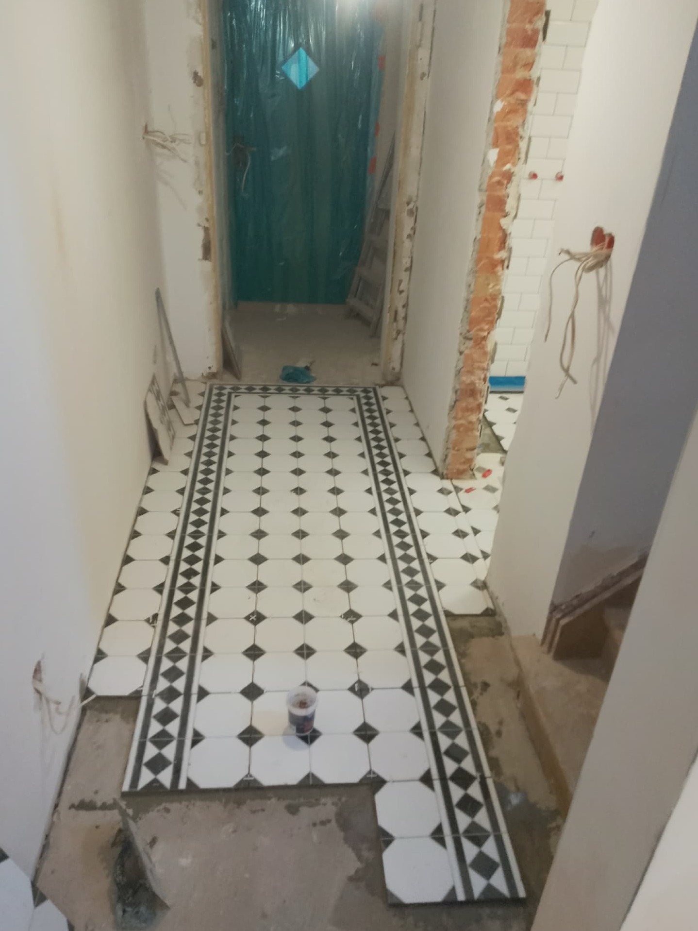 Układanie czarno-białej, wzorzystej podłogi w korytarzu. Widoczne niedokończone prace, surowe ściany i wystające kable. W tle zabezpieczone folią drzwi.