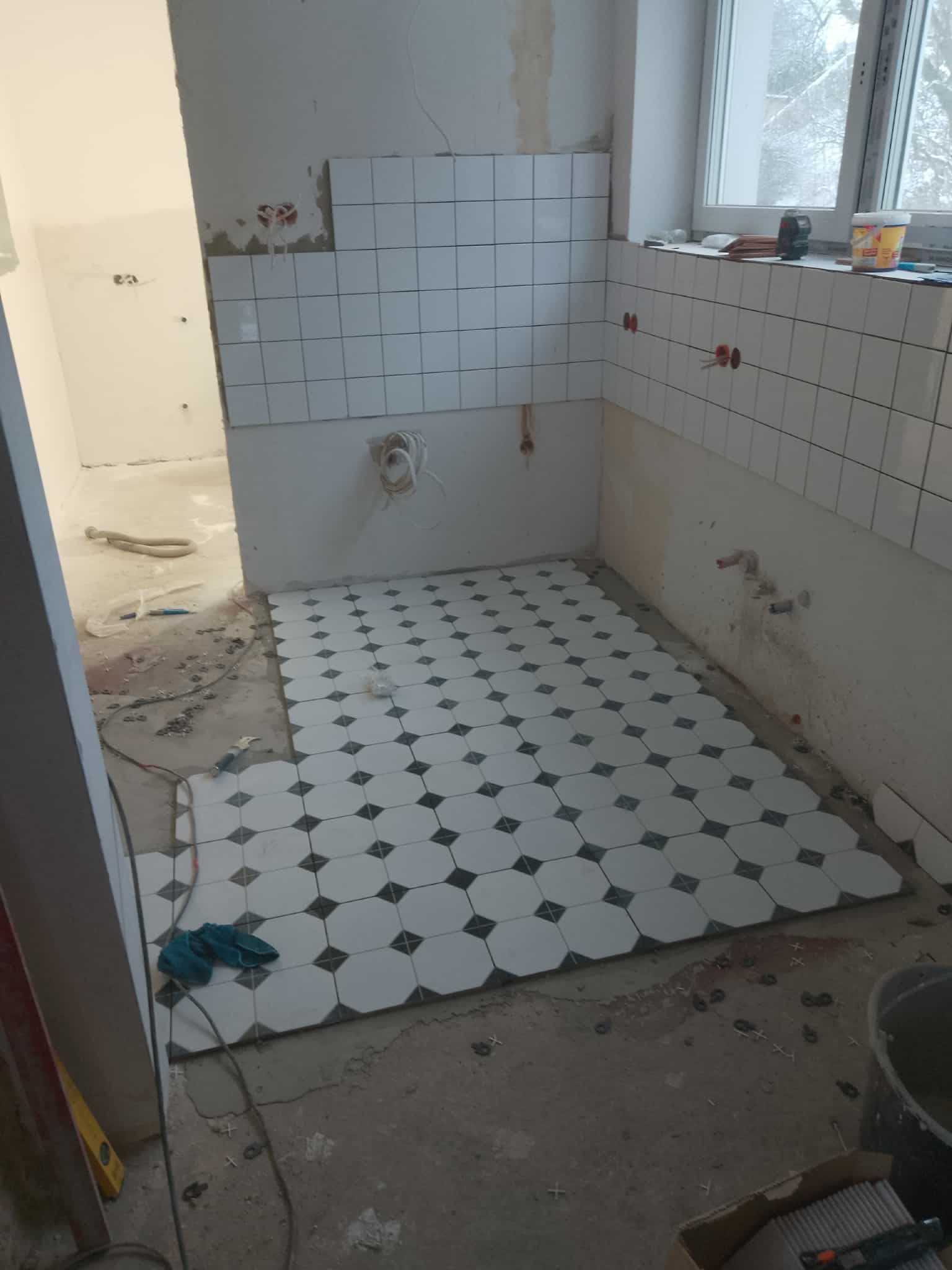 Układanie podłogi z płytek w łazience. Widoczne białe płytki z czarnymi akcentami. Częściowo ułożone płytki na ścianie. Prace w toku, widoczne narzędzia i materiały.