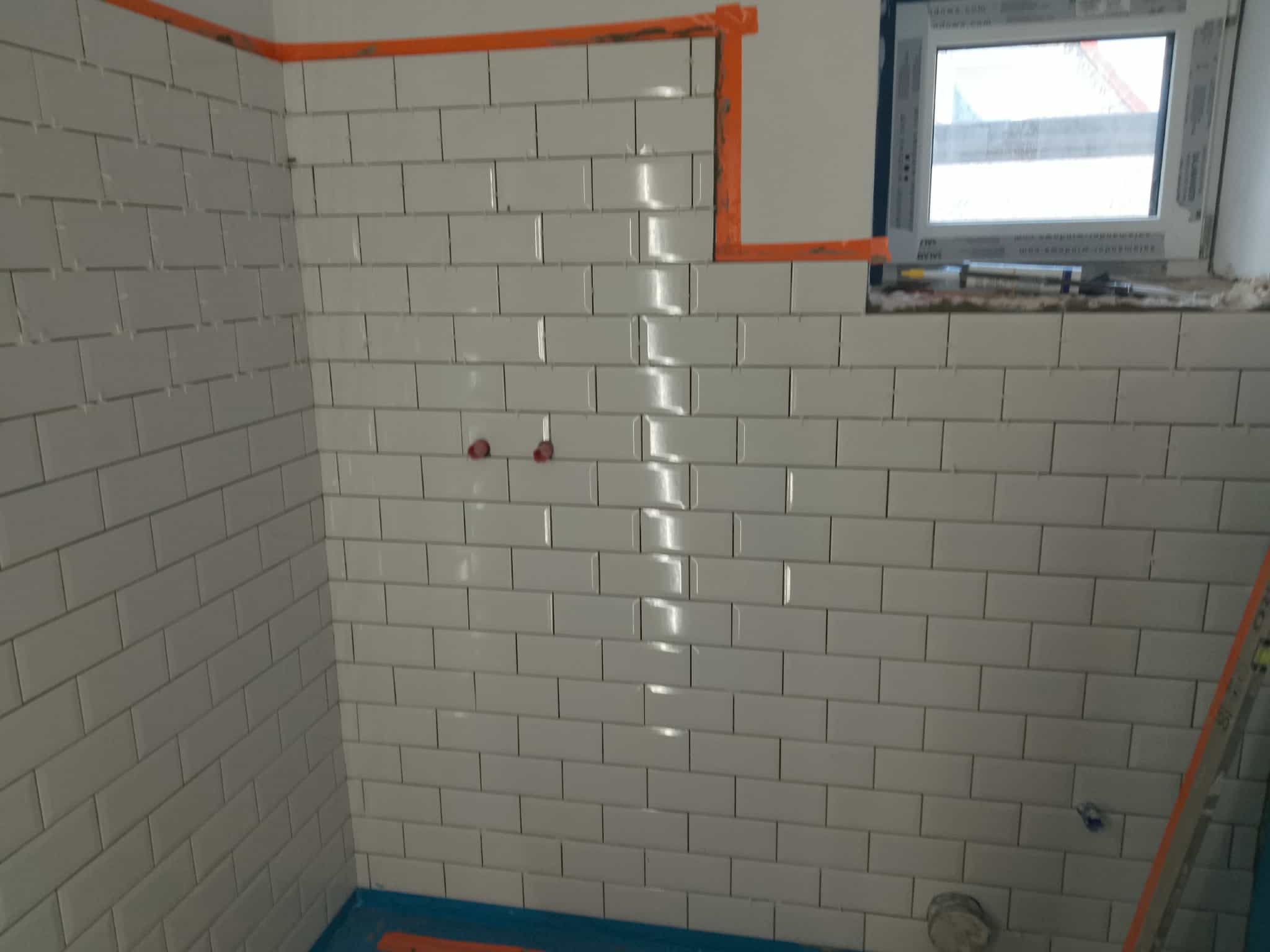 Ściana łazienki wyłożona białymi, błyszczącymi płytkami typu metro, z widocznymi otworami na instalacje i fragmentem okna w górnym rogu. Widoczne pomarańczowe taśmy zabezpieczające.