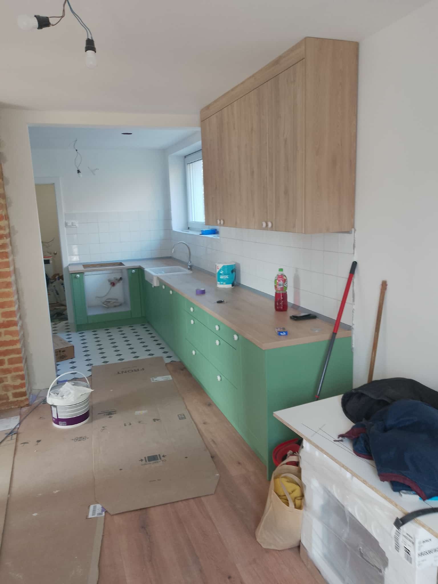 Montaż zielonej zabudowy kuchennej z drewnianym blatem i górnymi szafkami. Widoczne białe płytki na ścianie i podłoga zabezpieczona kartonem.
