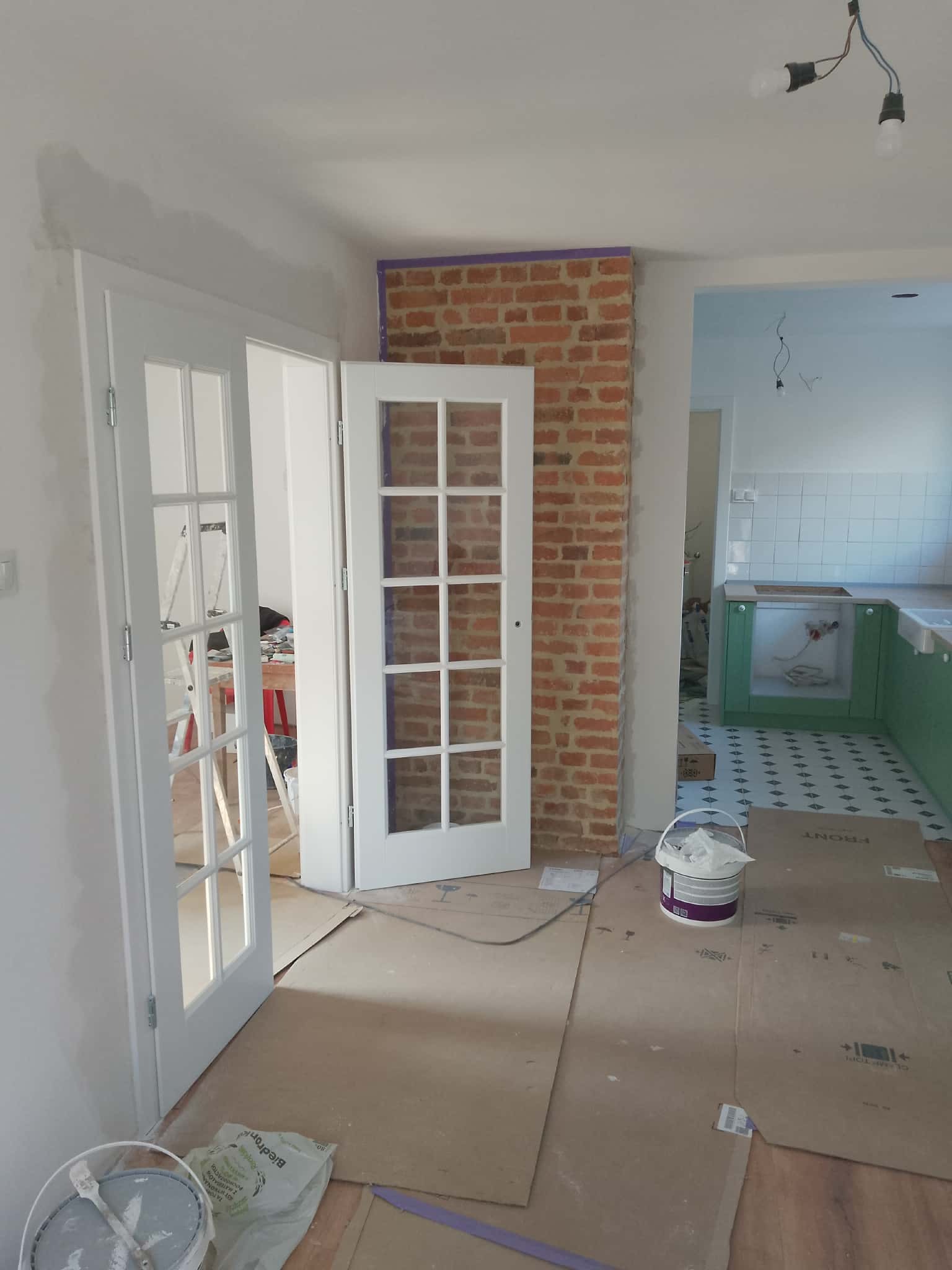 Wnętrze w trakcie prac: białe, przeszklone drzwi uchylne, fragment ściany z cegły zabezpieczony taśmą, podłoga wyłożona kartonem, widoczne narzędzia malarskie.