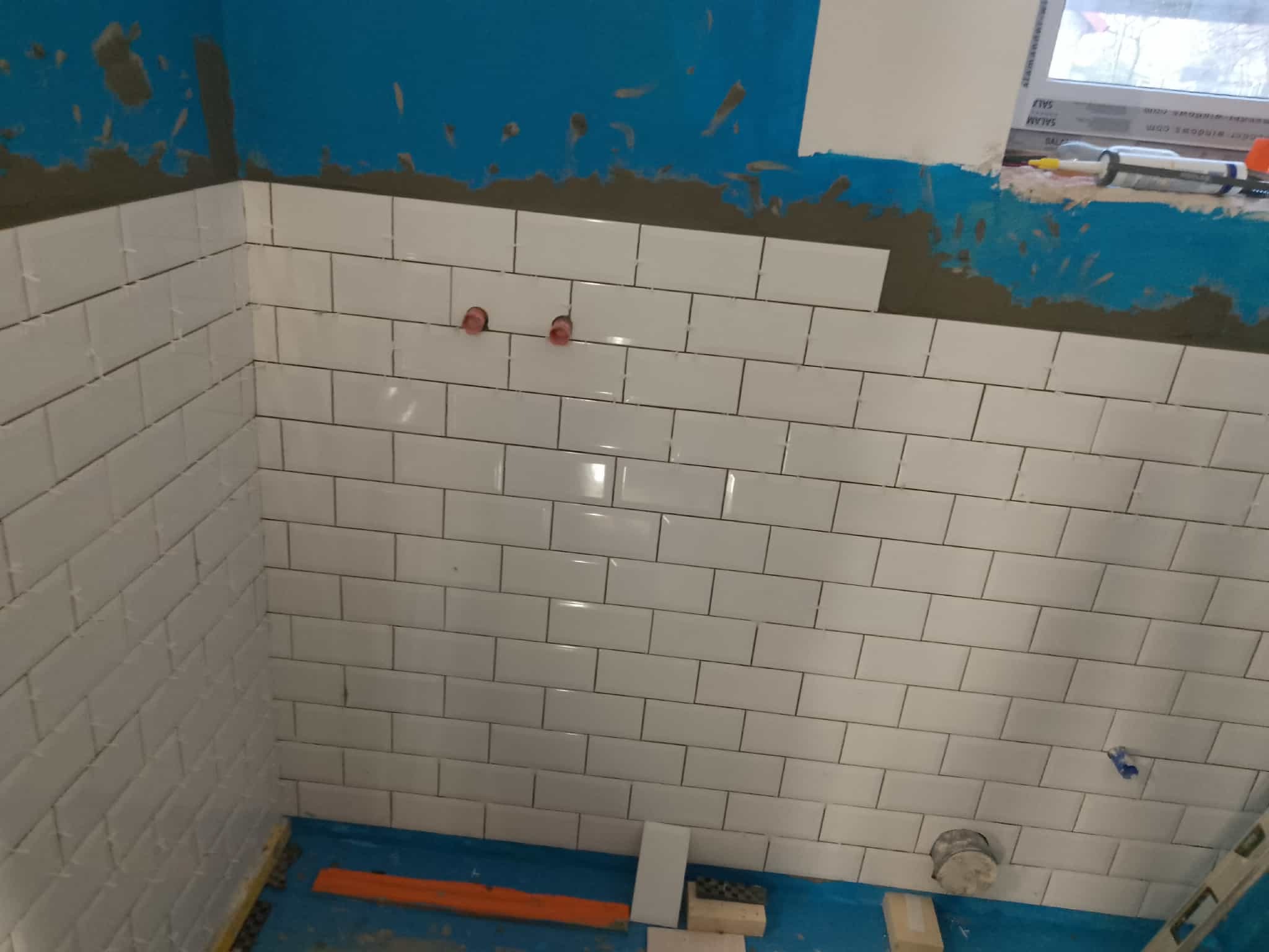 Białe, prostokątne płytki ułożone na ścianie łazienki, widoczne fragmenty niebieskiej ściany i narzędzia glazurnicze. Częściowo ukończona realizacja.