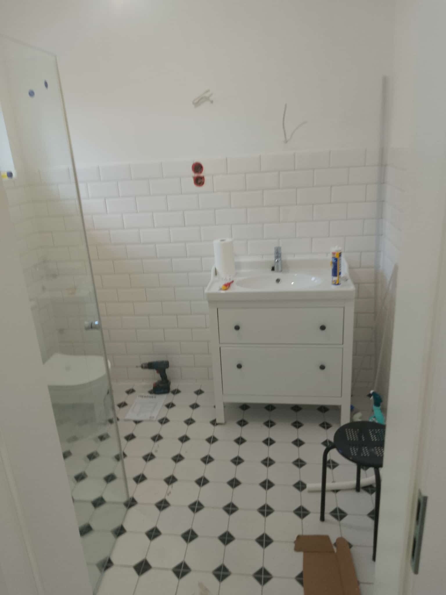 Biała łazienka po remoncie: umywalka z szafką, białe płytki metro, podłoga w czarno-białe wzory. W tle kabina prysznicowa i narzędzia.