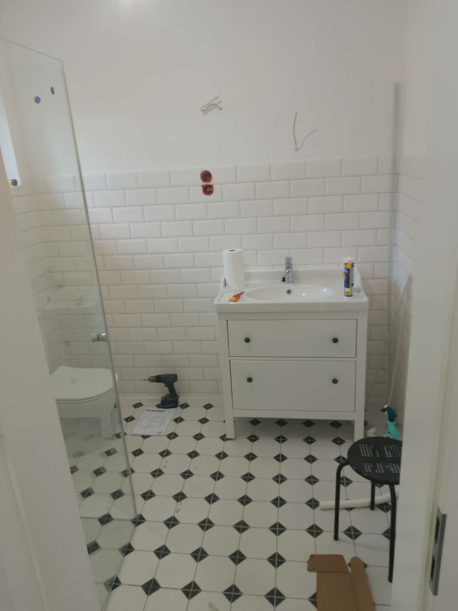 Biała łazienka z umywalką na szafce, białe płytki metro na ścianach, czarno-biała podłoga w karo. W tle kabina prysznicowa i narzędzia.