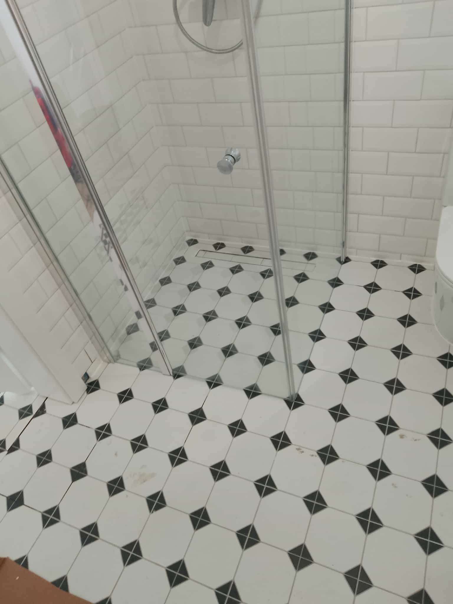 Wykończona łazienka z kabiną prysznicową i biało-czarną mozaiką na podłodze. Ściany wyłożone białymi, prostokątnymi płytkami. Widoczny fragment toalety.