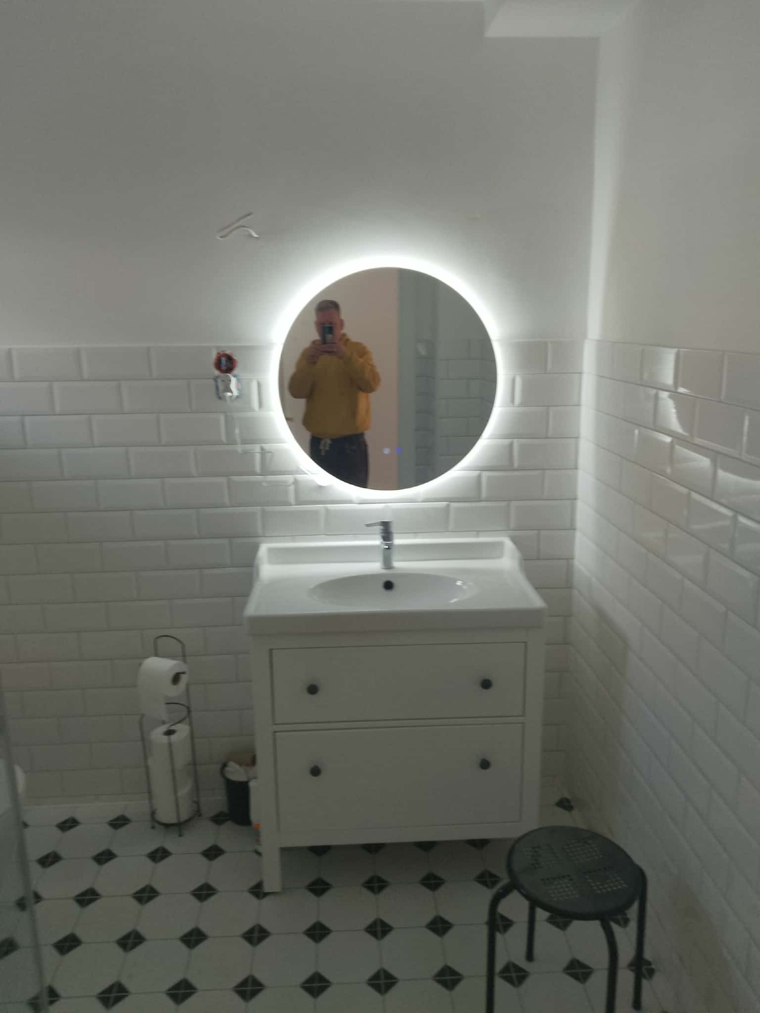 Biała łazienka z okrągłym lustrem LED, białą umywalką z szafką, białe płytki na ścianach i czarno-biała podłoga. W lustrze odbija się osoba robiąca zdjęcie.