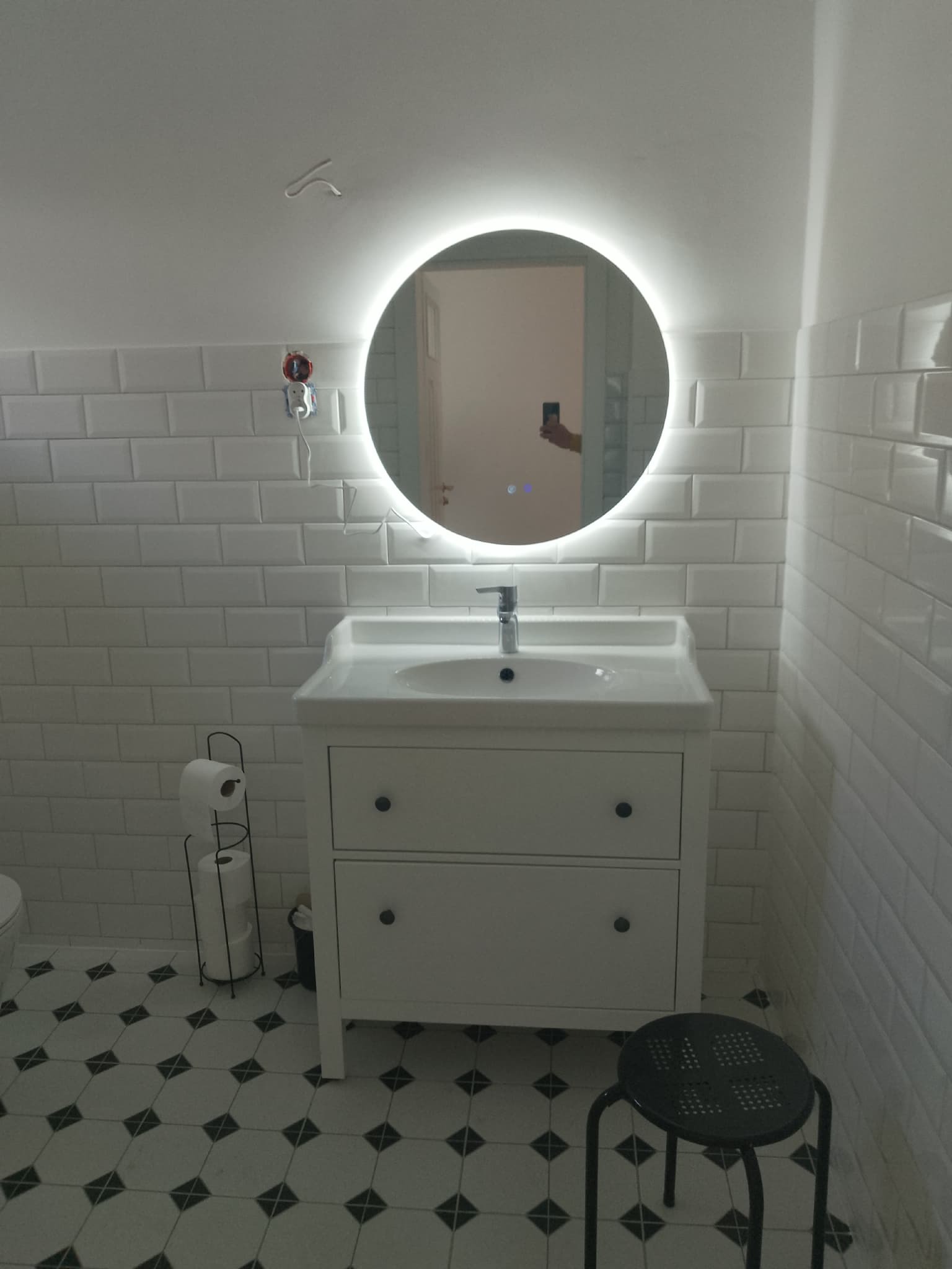 Biała łazienka z umywalką w szafce, okrągłym lustrem z podświetleniem LED i wzorzystą podłogą. Widoczne białe płytki imitujące cegłę i czarny stołek.