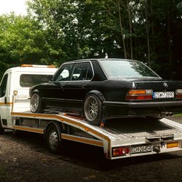 Przemysław Domański - Czarny, klasyczny BMW serii 5 (E28) na białej lawecie z pomarańczowym światłem ostrzegawczym, na tle zieleni drzew. Widoczny numer rejestracyjny.