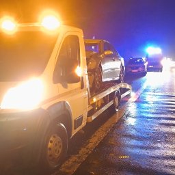 Przemysław Domański - Laweta z uszkodzonym autem na platformie, nocą na mokrej drodze. W tle światła innych pojazdów i odblaski na asfalcie. Widoczny numer rejestracyjny lawety.