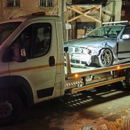 Przemysław Domański - Laweta z uszkodzonym srebrnym BMW na platformie, nocne ujęcie. W tle budynek i inne pojazdy, w tym furgonetka z reklamą firmy budowlanej.