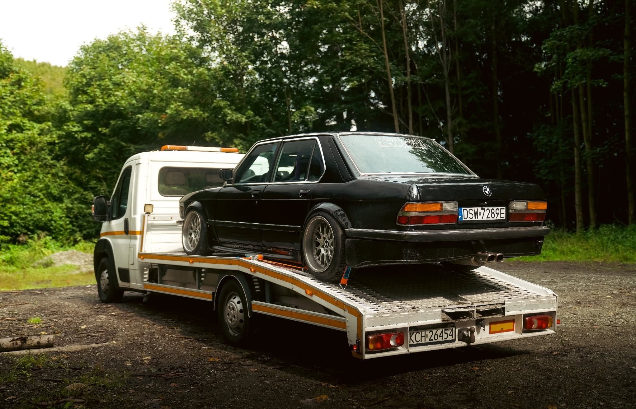 Czarna limuzyna BMW E28 na białej lawecie z pomarańczowymi pasami, widok z tyłu, tablica rejestracyjna DSW-7289E, las w tle.