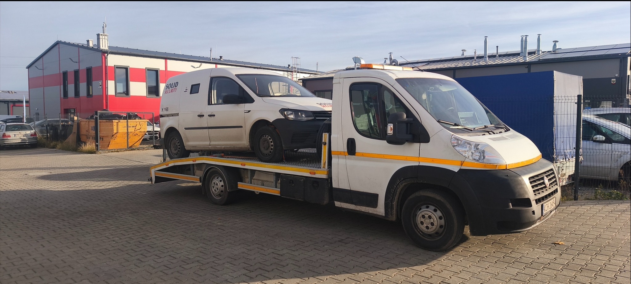 Laweta z białym vanem Solid Security na platformie, zaparkowana na brukowanym placu. W tle budynek z czerwono-białymi pasami i inne pojazdy.