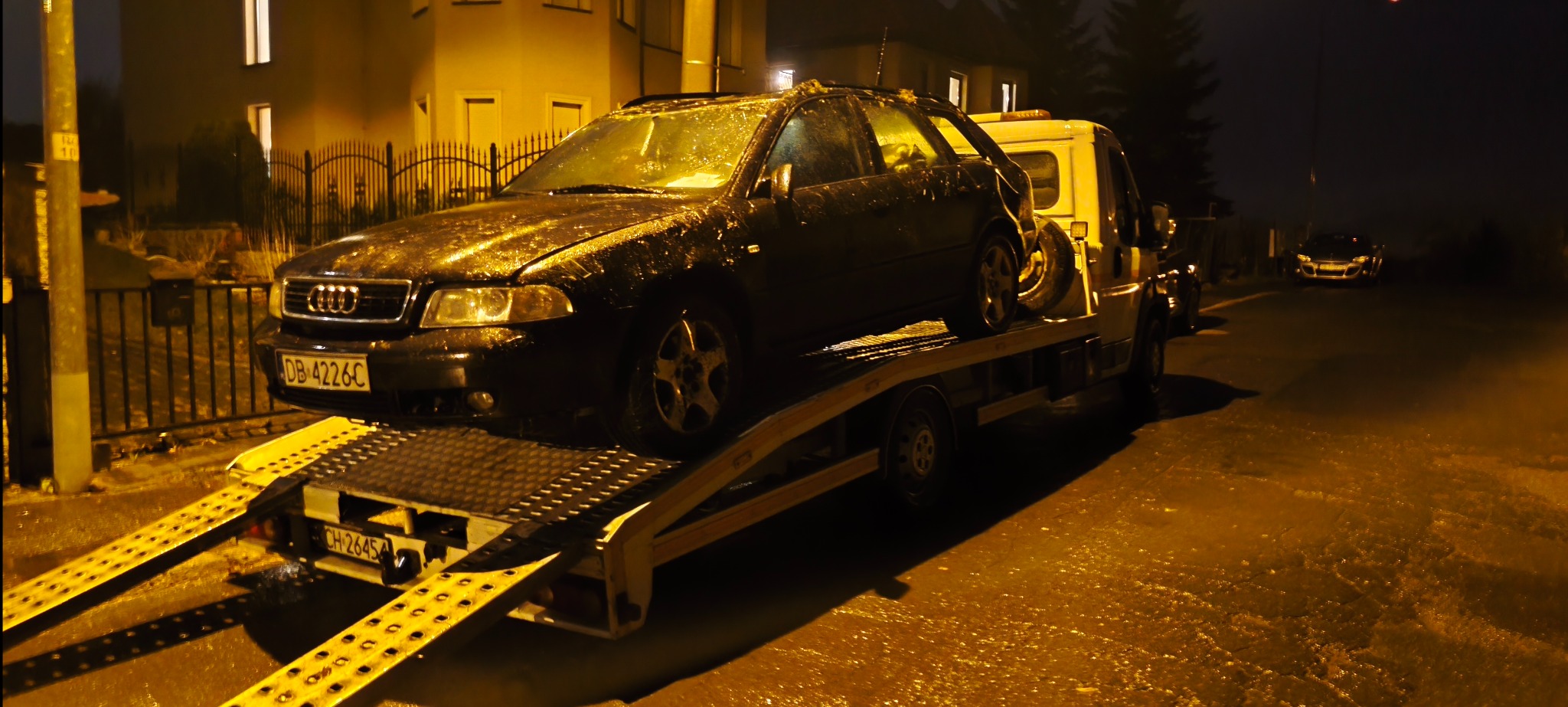 Czarny samochód Audi na żółtej lawecie w nocy, mokra nawierzchnia, oświetlenie uliczne. Widoczny numer rejestracyjny pojazdu i lawety.
