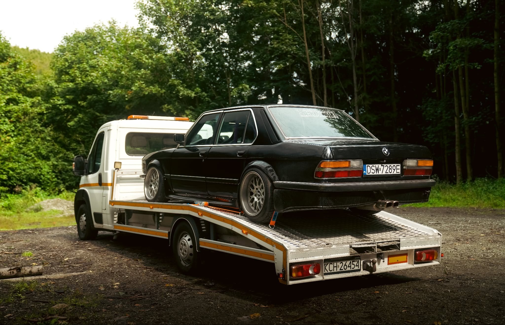 Czarny, klasyczny BMW serii 5 (E28) na białej lawecie z pomarańczowym światłem ostrzegawczym, na tle zieleni drzew. Widoczny numer rejestracyjny.