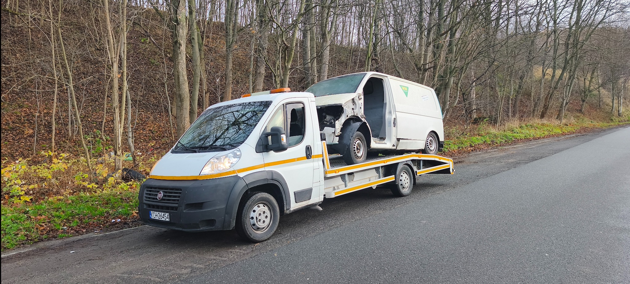 Laweta przewozi uszkodzony biały samochód dostawczy na tle jesiennego krajobrazu. Widoczny fragment drogi i drzewa bez liści.