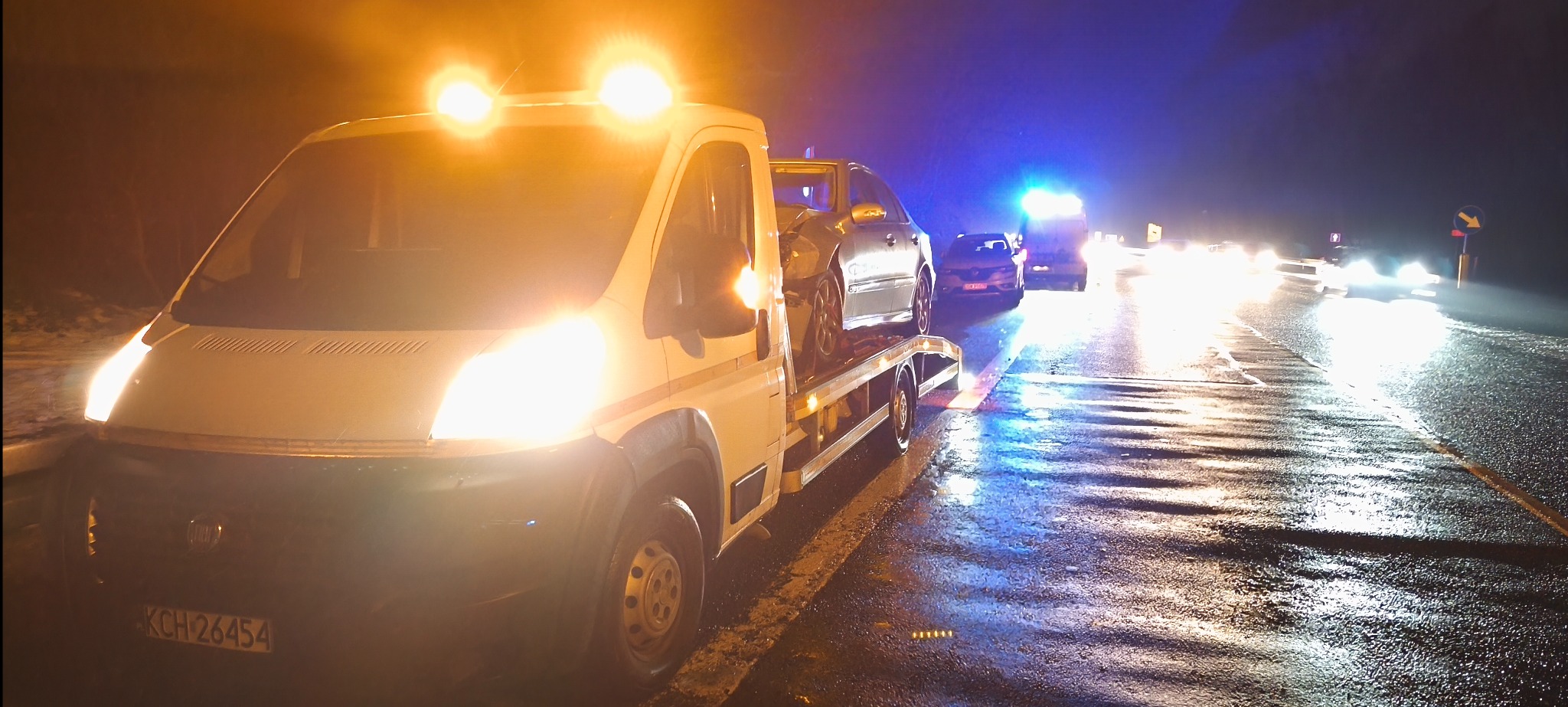 Laweta z uszkodzonym autem na platformie, nocą na mokrej drodze. W tle światła innych pojazdów i odblaski na asfalcie. Widoczny numer rejestracyjny lawety.