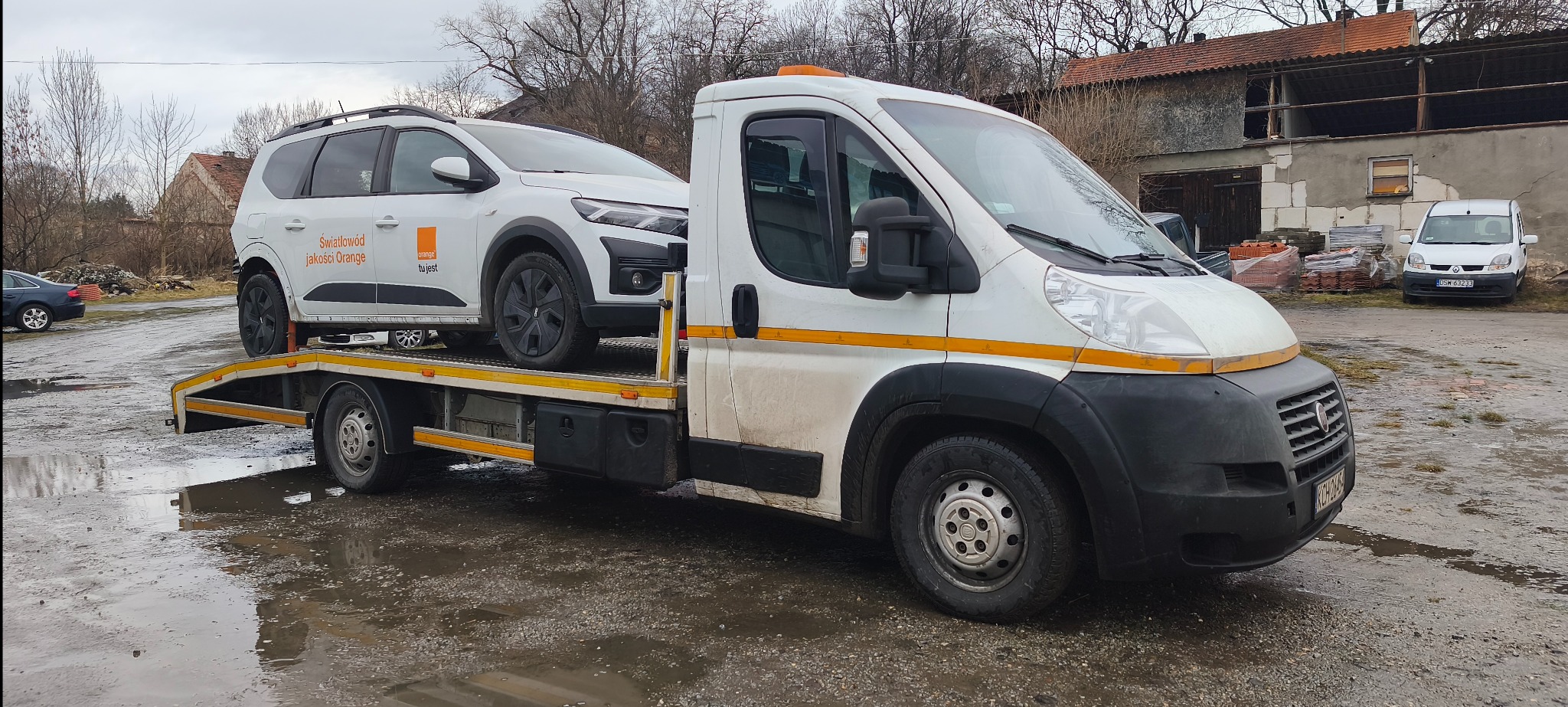 Laweta z białym samochodem marki Dacia z logo Orange na platformie, na tle zabudowań i kałuż. Transport pojazdu dla firmy telekomunikacyjnej.