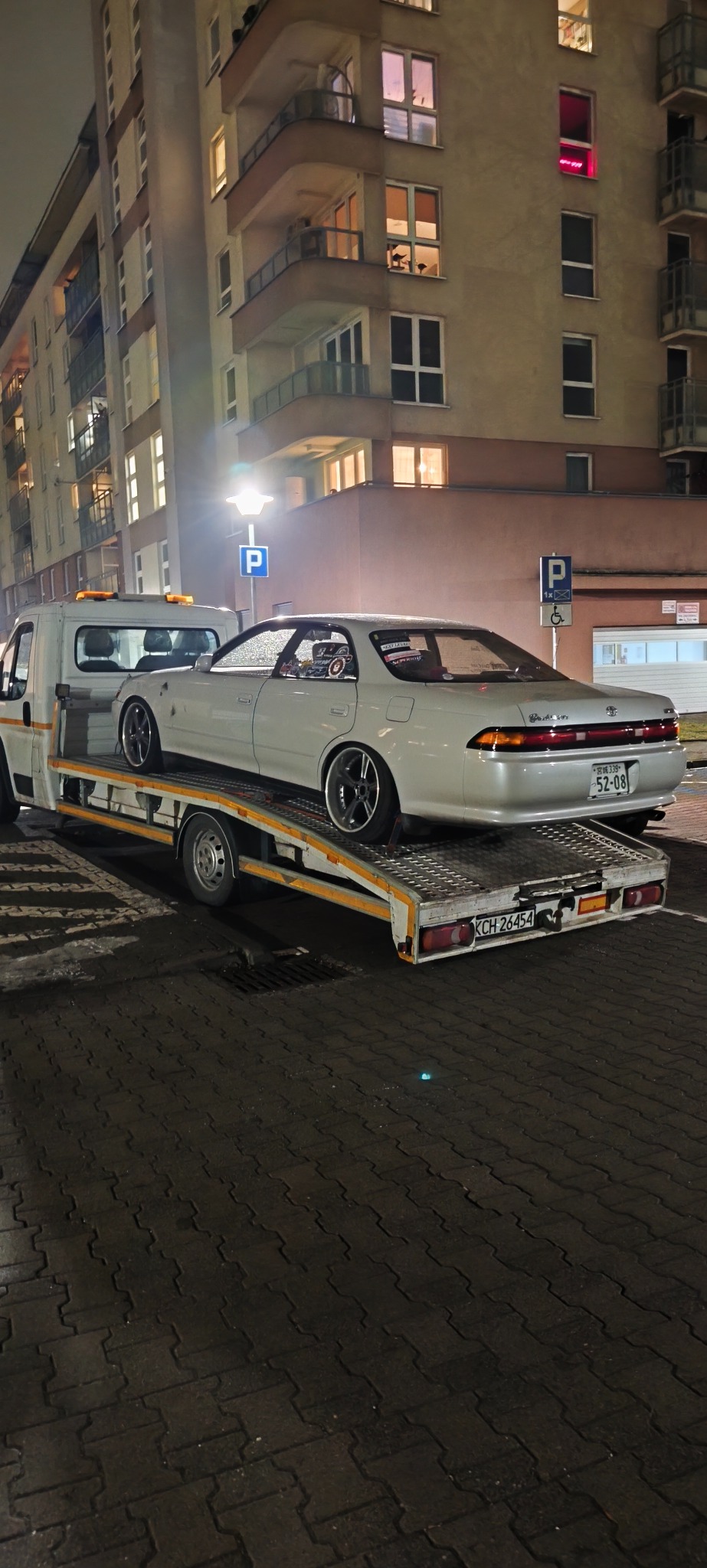 Laweta z białym sedanem Toyota Mark II na tle miejskiej zabudowy nocą. Widoczne naklejki na tylnej szybie auta i tablica rejestracyjna.