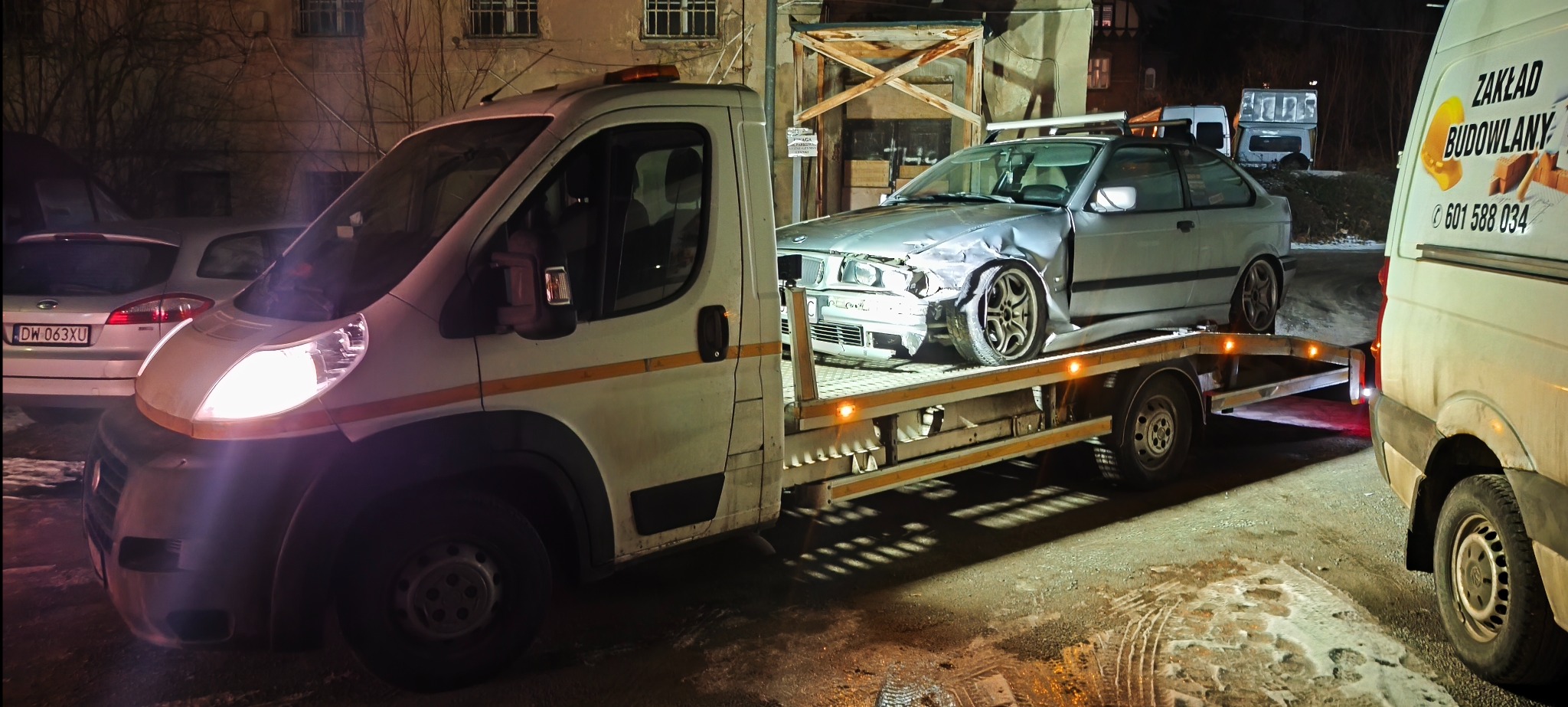 Laweta z uszkodzonym srebrnym BMW na platformie, nocne ujęcie. W tle budynek i inne pojazdy, w tym furgonetka z reklamą firmy budowlanej.
