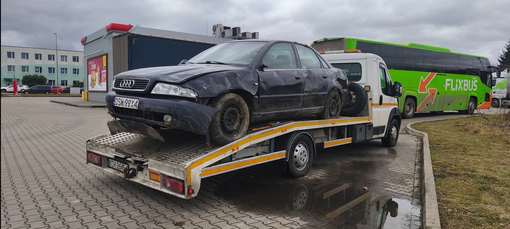 Czarny, uszkodzony samochód Audi A4 na lawecie, z widocznym autobusem Flixbus w tle. Tablica rejestracyjna DSW 99Y4. Widoczne uszkodzenia karoserii.