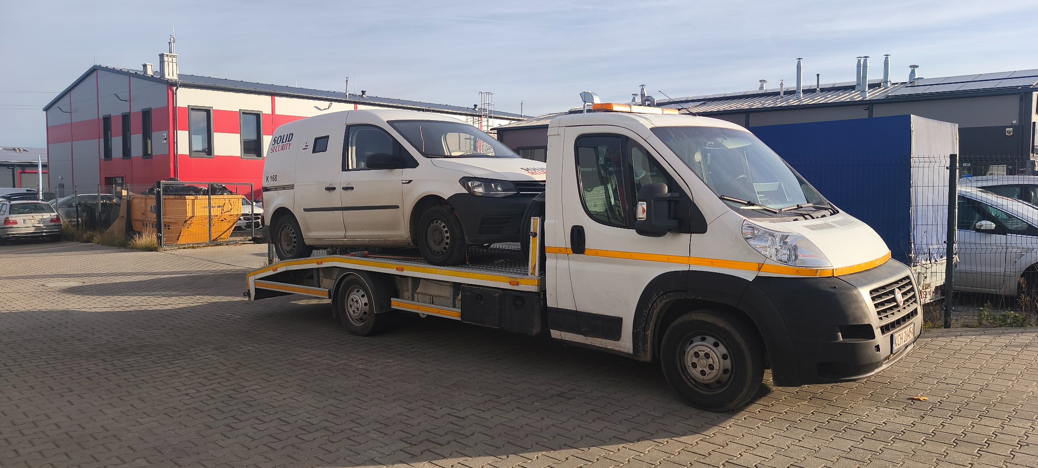 Laweta z białym samochodem dostawczym Solid Security na platformie, zaparkowana na brukowanym placu, budynek w tle. Żółte pasy na lawecie.