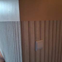 Michał Db - Wykończona ściana z panelami dekoracyjnymi w odcieniach beżu i brązu, z widocznym włącznikiem światła. W tle narzędzia pracy, sugerujące świeży montaż.