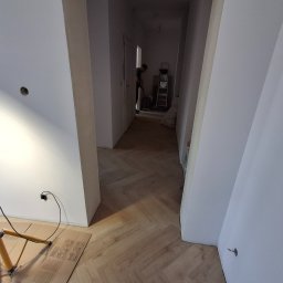 Michał Db - Jasne panele ułożone w jodełkę w korytarzu, widoczna lampa budowlana i pracownik w oddali. Świeżo pomalowane ściany i przygotowane podłoże.