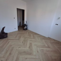 Michał Db - Jasne panele ułożone w jodełkę na podłodze w pustym pokoju. Widoczny odkurzacz, wiadro i torba. Świeżo wykończona podłoga w nowym mieszkaniu.