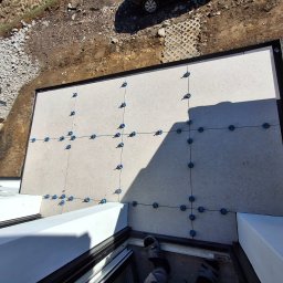 Michał Db - Jasne płytki tarasowe z czarną ramą, wyrównane niebieskimi krzyżykami dystansowymi, widok z góry na częściowo wykończony taras.