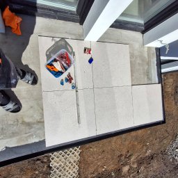 Michał Db - Jasne płytki tarasowe układane przy oknie, widok z góry. Częściowo widoczna osoba w roboczym ubraniu i narzędzia glazurnicze. Prace wykończeniowe na zewnątrz budynku.