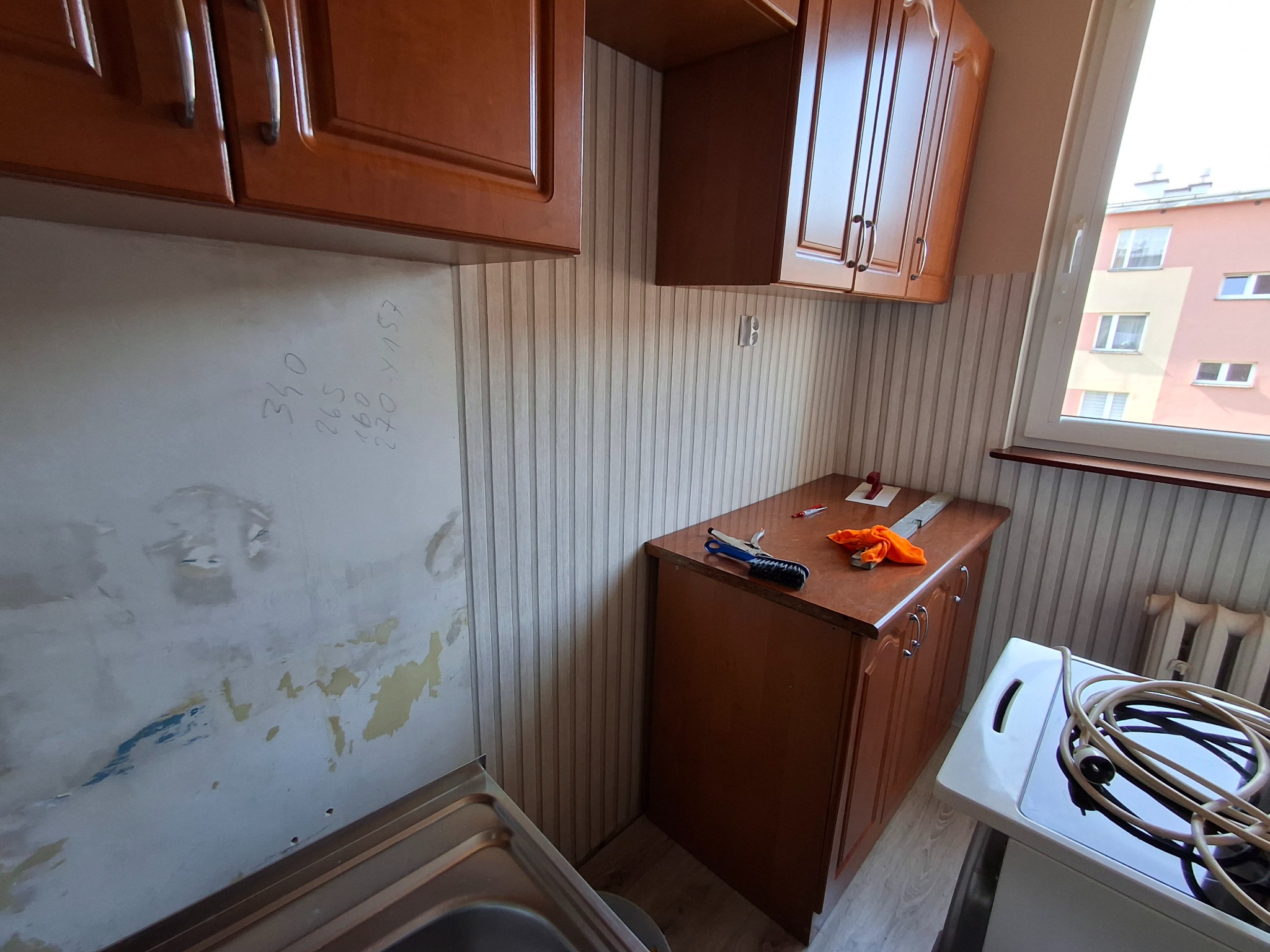 Fragment kuchni w trakcie prac przygotowawczych: ściana ze śladami po usuniętych płytkach, szafki wiszące, narzędzia na blacie. Widoczne instalacje.