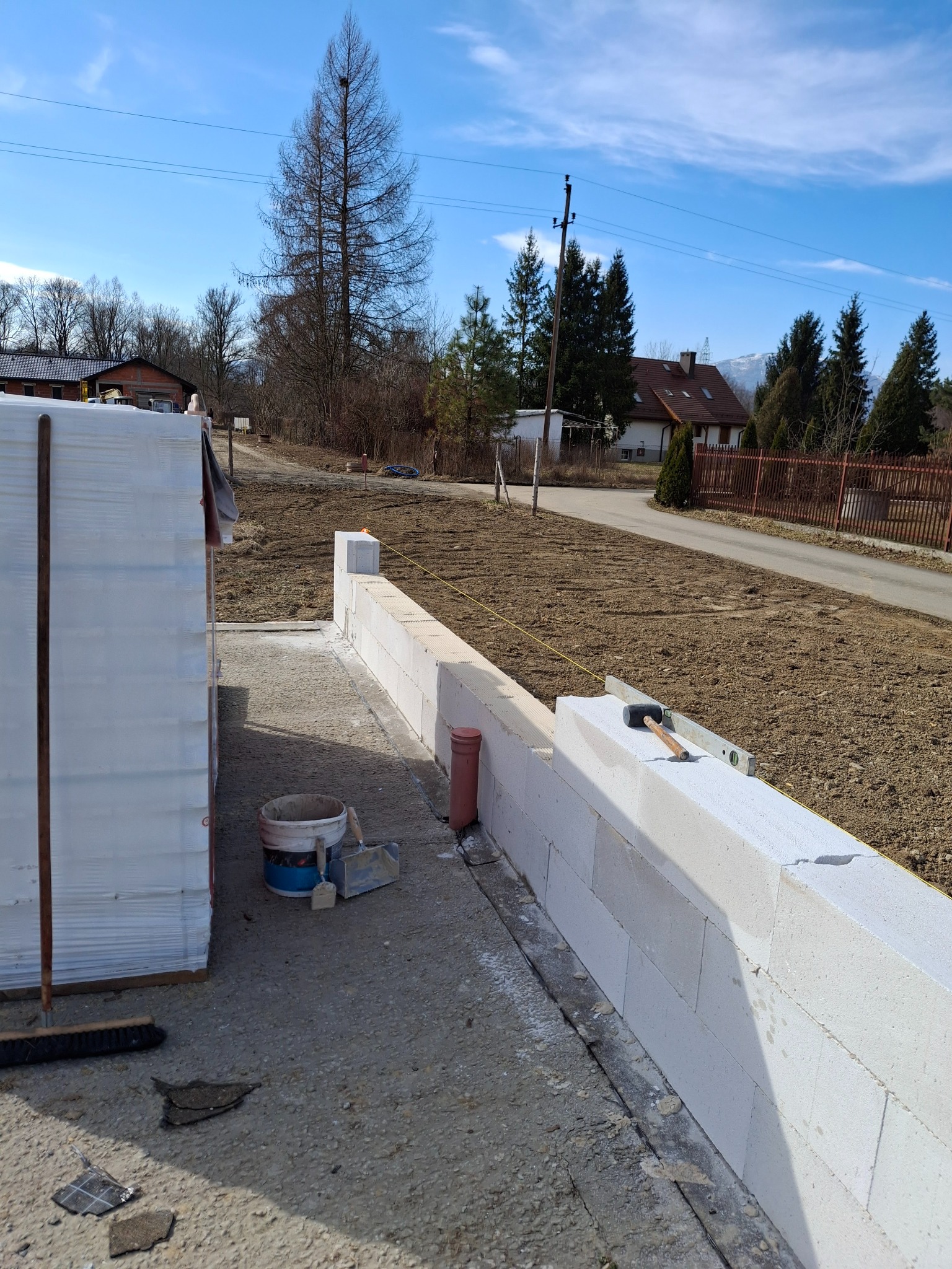 Wznoszenie muru z białych bloczków betonowych na zewnątrz. Widoczne narzędzia: poziomica, młotek, wiadro z zaprawą. W tle krajobraz z drzewami i zabudowaniami.