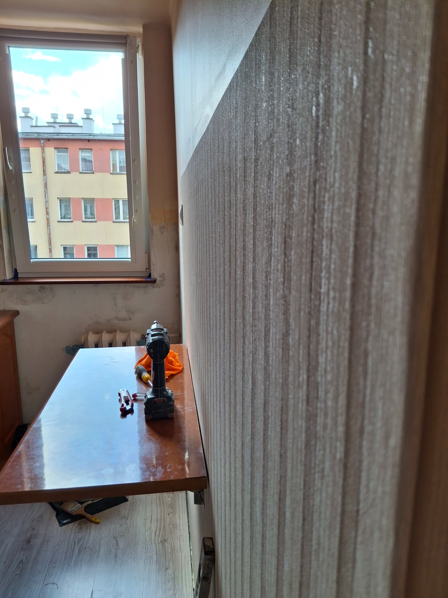 Ściana z tapetą w pionowe pasy, stół z wiertarką i narzędziami, okno z widokiem na budynek. Przygotowania do prac wykończeniowych w pomieszczeniu.