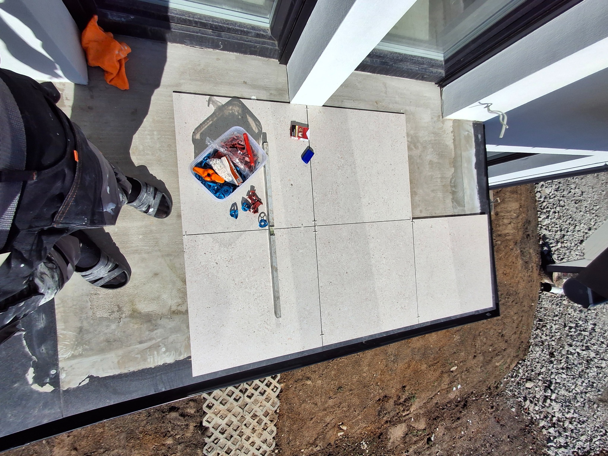 Jasne płytki tarasowe układane przy oknie, widok z góry. Częściowo widoczna osoba w roboczym ubraniu i narzędzia glazurnicze. Prace wykończeniowe na zewnątrz budynku.