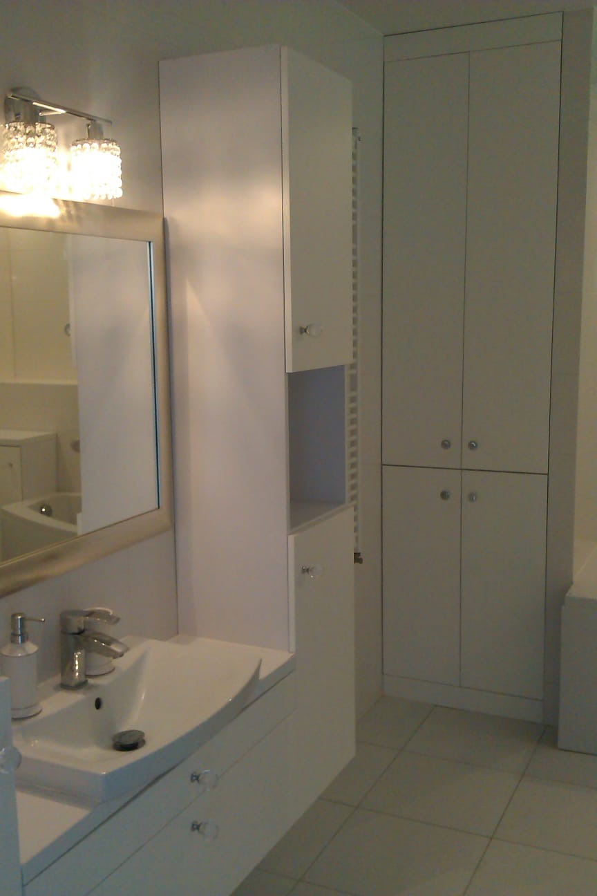 Jasna łazienka po remoncie: białe szafki z kryształowymi gałkami, umywalka z baterią, lustro z oświetleniem. Minimalistyczny design, płytki podłogowe w odcieniach szarości.