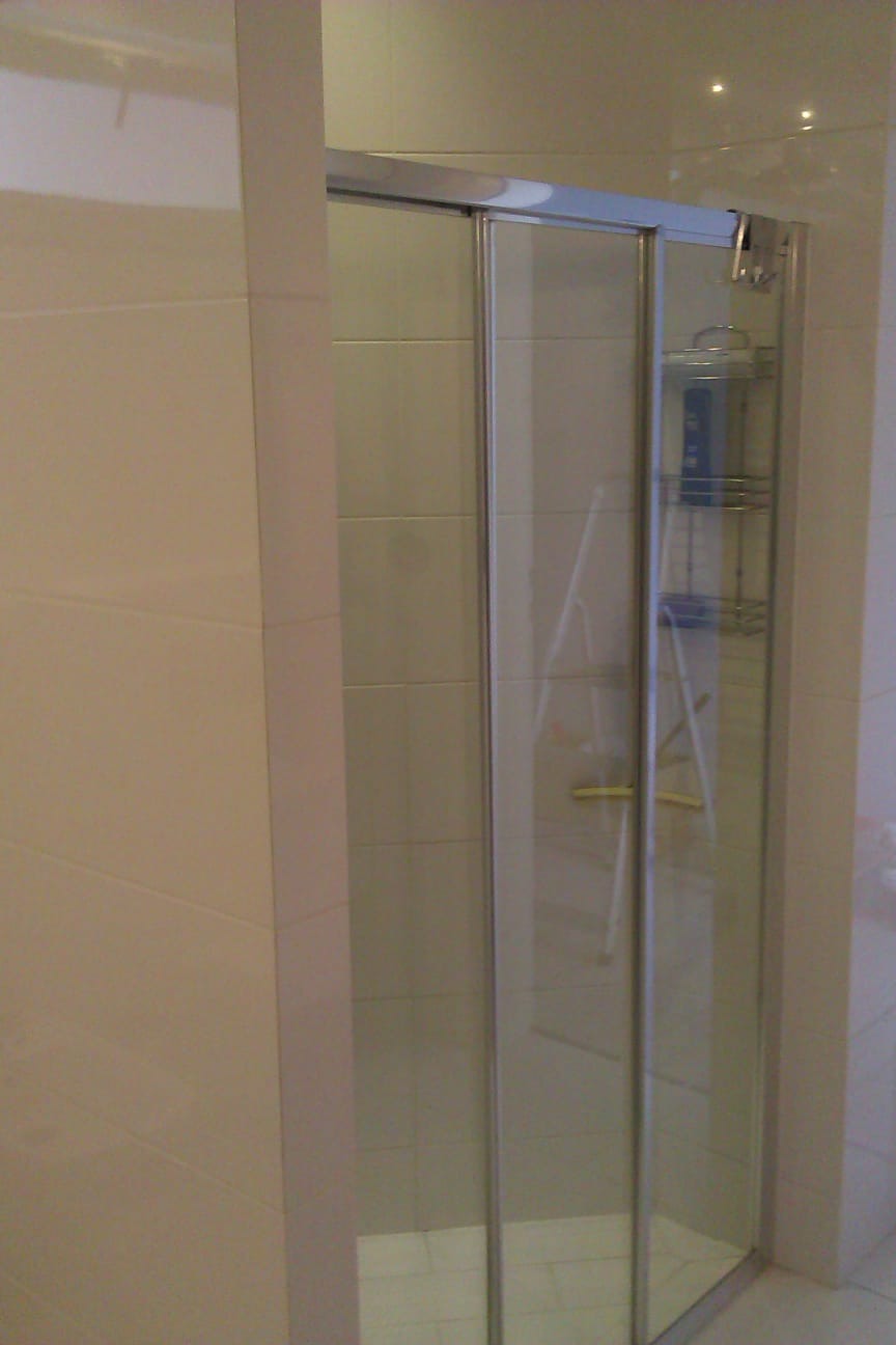 Zamknięta kabina prysznicowa z przesuwnymi drzwiami, wykończona beżowymi płytkami. Widoczna półka na kosmetyki wewnątrz kabiny i drabina oparta o ścianę.