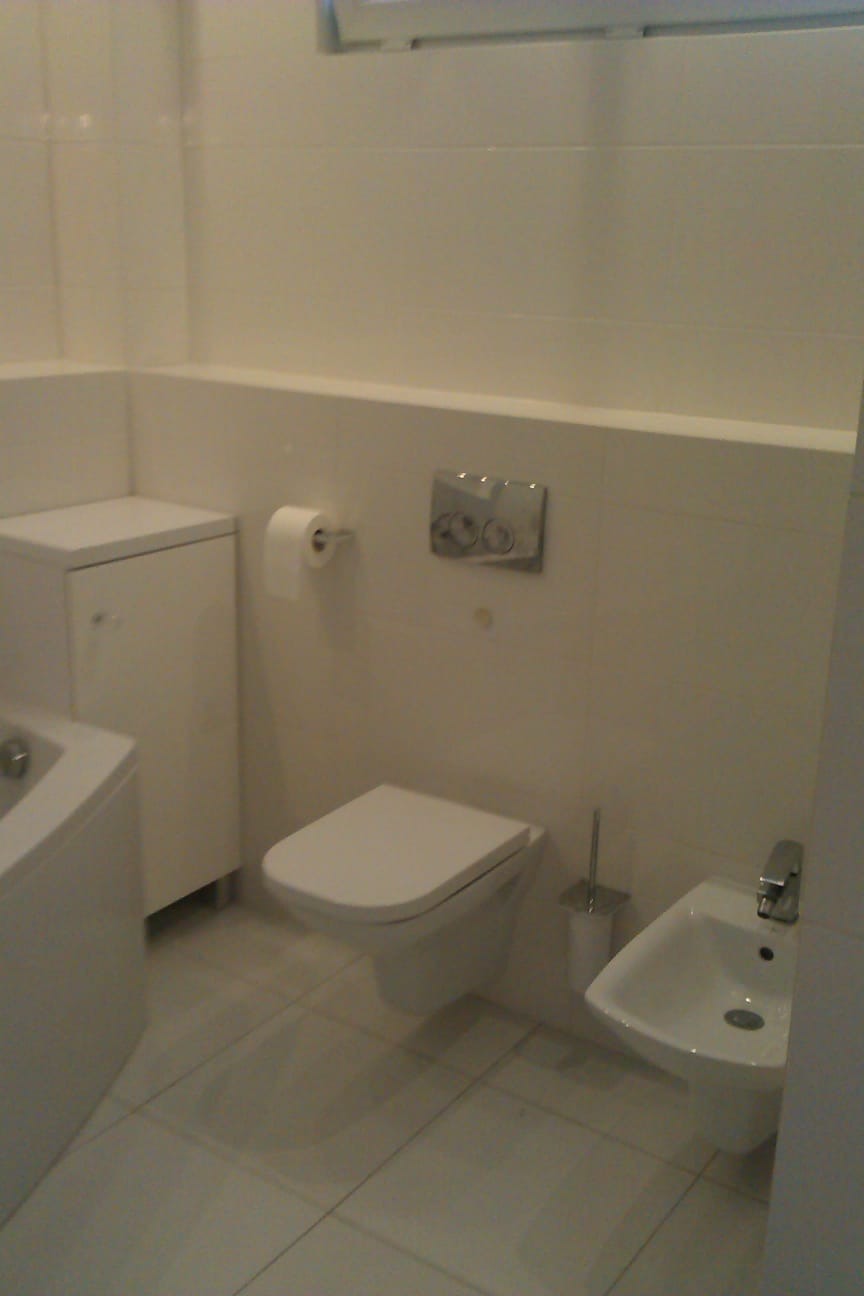 Biała, minimalistyczna łazienka z podwieszaną toaletą, bidetem i szafką narożną. Uwagę zwraca chromowany przycisk spłuczki i delikatne oświetlenie.